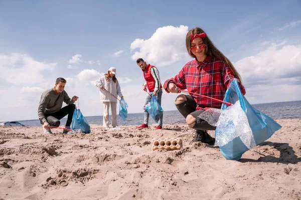 Giovane donna sorridente tenendo sacchetto della spazzatura e raccogliendo spazzatura con afferratore sulla sabbia vicino al gruppo di volontari — Foto stock