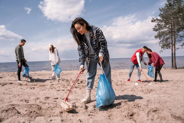 Sorrindo jovem segurando saco de lixo e coletando lixo na areia perto do grupo de voluntários — Fotografia de Stock