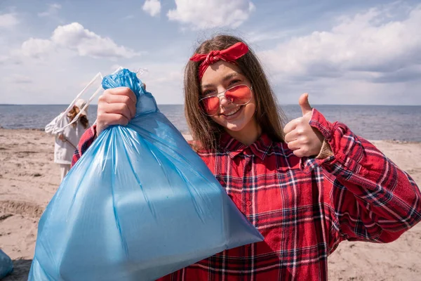 Mujer joven feliz en gafas de sol sosteniendo la bolsa de basura y mostrando el pulgar hacia arriba, concepto de ecología - foto de stock