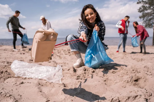 Mujer joven borrosa recogiendo basura con agarrador cerca de grupo de voluntarios - foto de stock