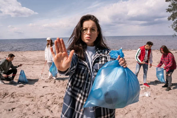 Mujer joven mostrando gesto de stop cerca de grupo borroso de voluntarios recogiendo basura, concepto de ecología - foto de stock