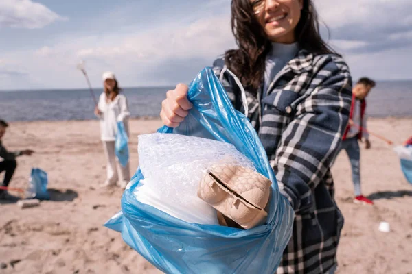 Cropped view of smiling young woman holding trash bag with rubbish near group of volunteers — Stock Photo