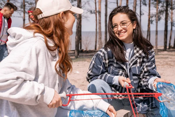 Mujeres jóvenes felices con agarradores recogiendo basura fuera - foto de stock