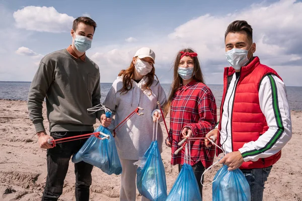 Jóvenes voluntarios en máscaras médicas sosteniendo bolsas de basura y agarradores fuera, recoger el concepto de basura - foto de stock