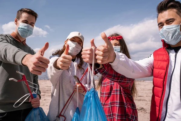 Jóvenes voluntarios con máscaras médicas sosteniendo bolsas de basura y agarradores mientras muestran los pulgares hacia arriba, recogen el concepto de basura - foto de stock