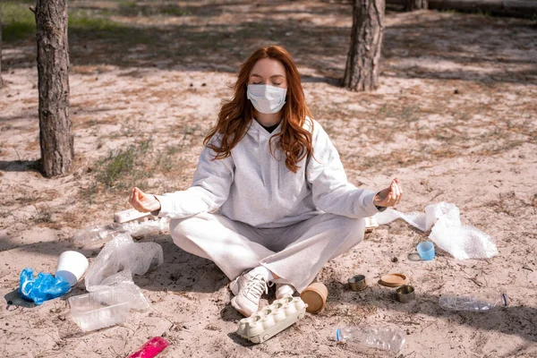 Young woman in medical mask meditating near trash on ground — Stock Photo