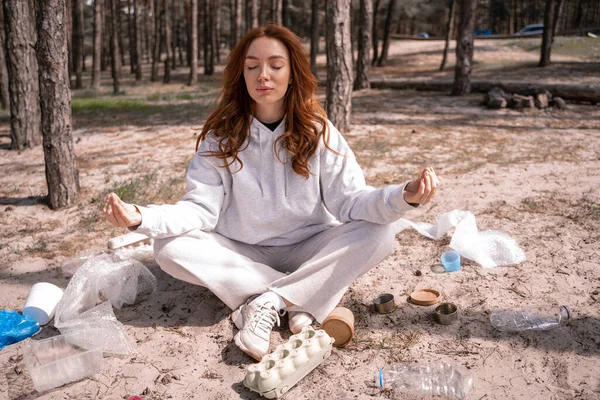 Jovem com olhos fechados meditando perto do lixo no chão — Fotografia de Stock