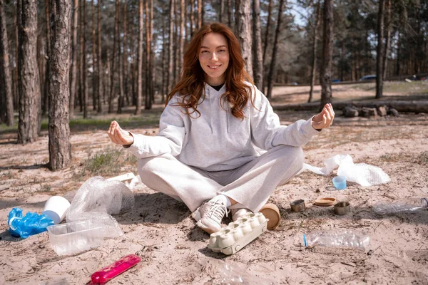 Happy young woman with crossed legs sitting and meditating near trash on ground — Stock Photo