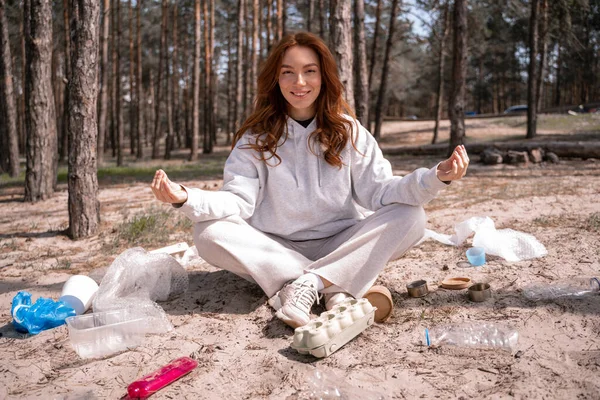 Feliz joven con las piernas cruzadas sentado y meditando cerca de la basura en el suelo - foto de stock