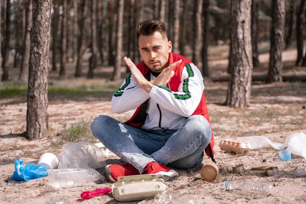 Man with crossed legs sitting and showing no sign near trash on ground — Stock Photo