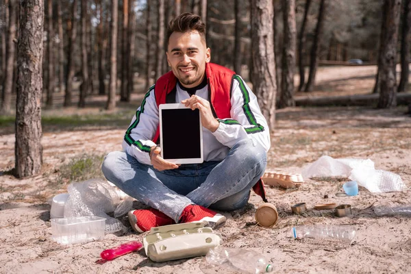 Hombre feliz con las piernas cruzadas sentado y sosteniendo tableta digital con pantalla en blanco cerca de la basura en el suelo - foto de stock