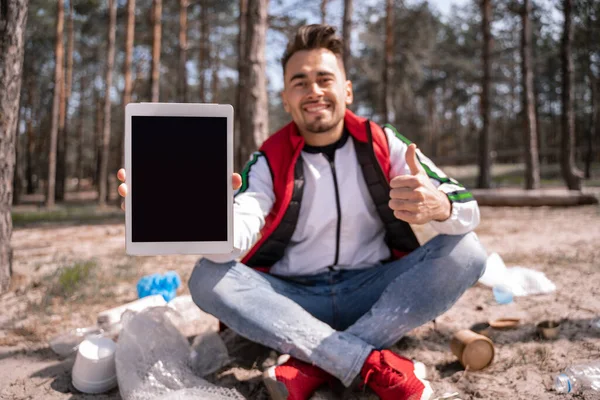 Happy man with crossed legs holding digital tablet with blank screen and showing thumb up near trash on ground — Stock Photo