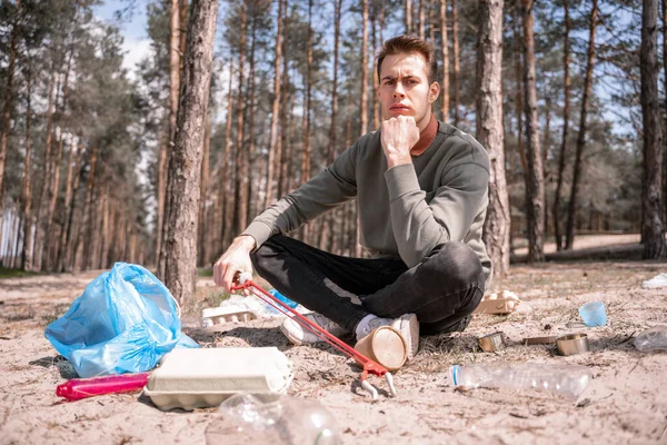 Hombre pensativo con las piernas cruzadas sentado cerca de la basura en el suelo en el bosque - foto de stock