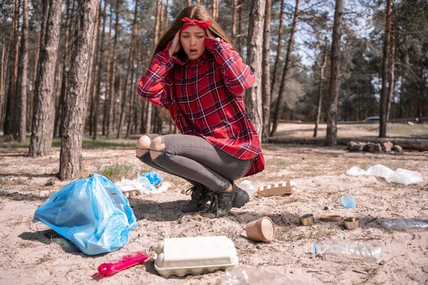 Шокована молода жінка дивиться на сміття біля сміттєвого мішка в лісі — стокове фото
