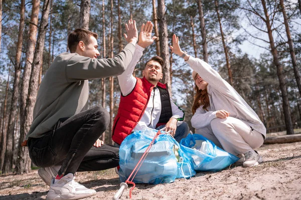 Volontari allegri dando alto cinque vicino sacchi della spazzatura nella foresta — Foto stock