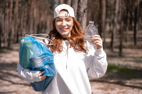 Щаслива молода жінка тримає синій мішок сміття і пластикову пляшку в лісі — стокове фото