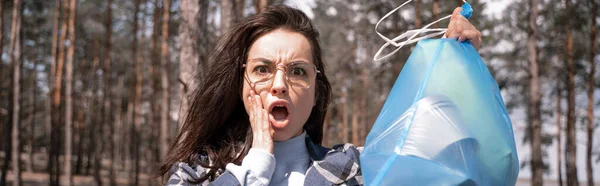 Шокированная молодая женщина держит синий мешок для мусора в лесу, баннер — стоковое фото