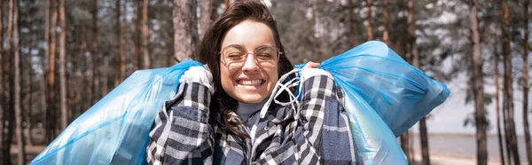 Allegra giovane donna in occhiali che tiene borse blu spazzatura nella foresta, banner — Foto stock