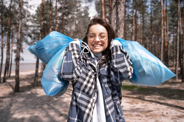 Alegre joven en gafas sosteniendo bolsas de basura azul en el bosque - foto de stock