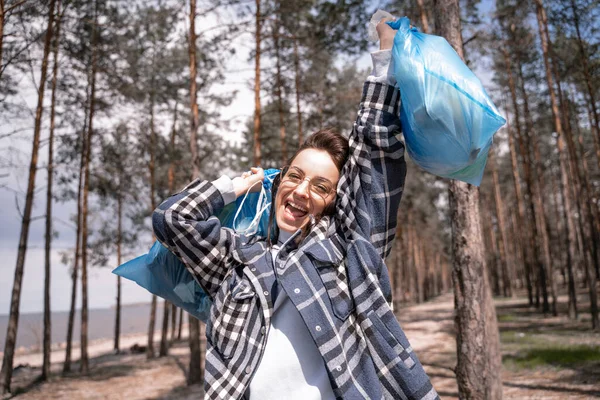 Збуджена молода жінка в окулярах тримає сині мішки для сміття в лісі — стокове фото