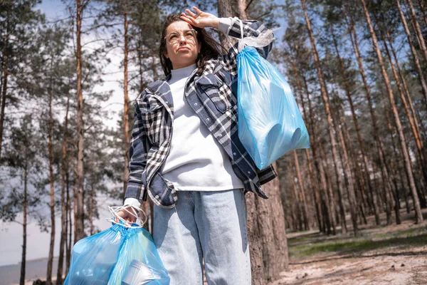 Displeased young woman in glasses holding blue trash bags in forest — Stock Photo