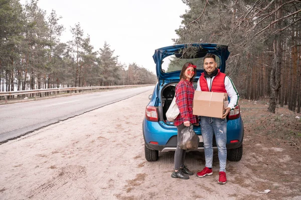 Happy woman in sunglasses holding trash bag while man standing with carton box near modern car in forest — Stock Photo