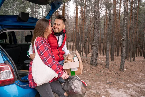 Женщина в солнцезащитных очках держит мусорный мешок, а счастливый мужчина стоит с коробкой рядом с машиной в лесу — стоковое фото