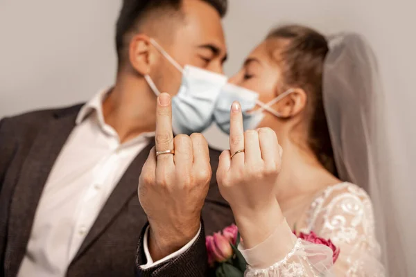 Blurred newlyweds in protective masks kissing while showing wedding rings isolated on grey — Stock Photo