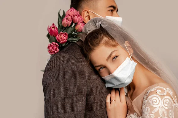 Young bride in medical mask leaning on chest of groom and looking at camera isolated on grey — Stock Photo