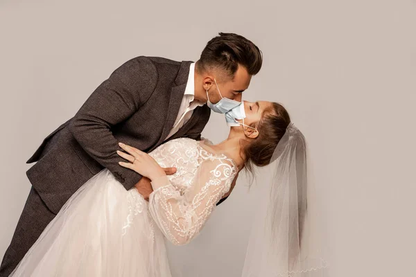 Чоловік в медичних масках цілує елегантну наречену ізольовано на сірому з бузковим відтінком — стокове фото