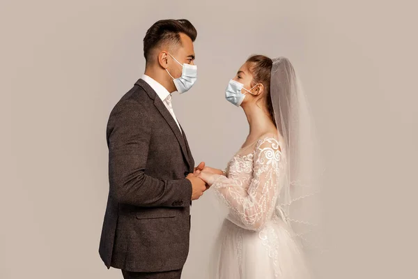 Side view of groom and bride in medical masks holding hands isolated on grey with lilac shade - foto de stock