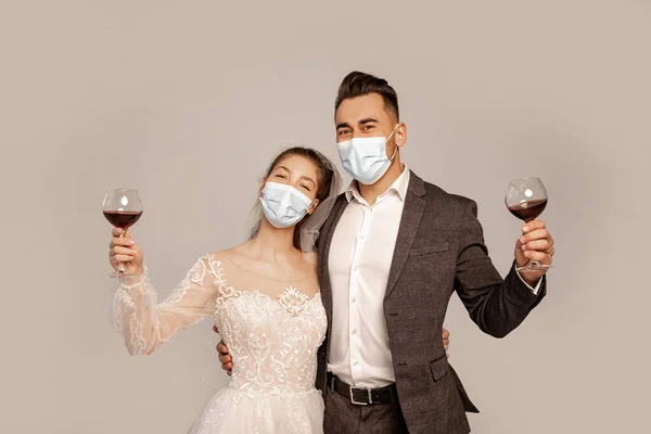 Young newlyweds in medical masks holding glasses with red wine isolated on grey — Stock Photo