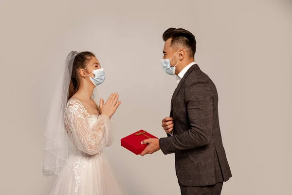 Side view of man in medical mask presenting gift box to elegant bride isolated on grey - foto de stock