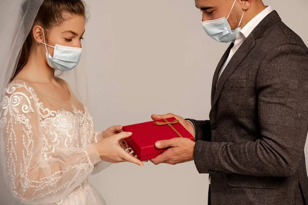 Mann in medizinischer Maske präsentiert Geschenkbox für junge Braut isoliert auf grau — Stockfoto