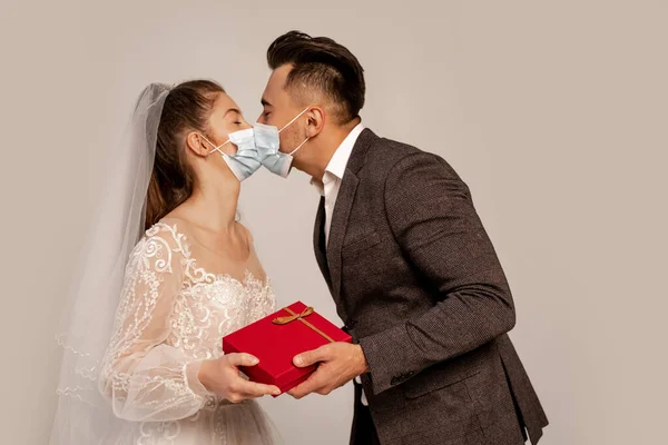 Groom in medical mask kissing bride while presenting gift isolated on grey — Photo de stock