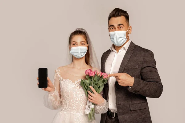 Man in medical mask pointing at smartphone in hand of bride isolated on grey — Photo de stock