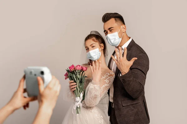 Newlyweds in medical masks showing wedding rings near blurred photographer isolated on grey - foto de stock