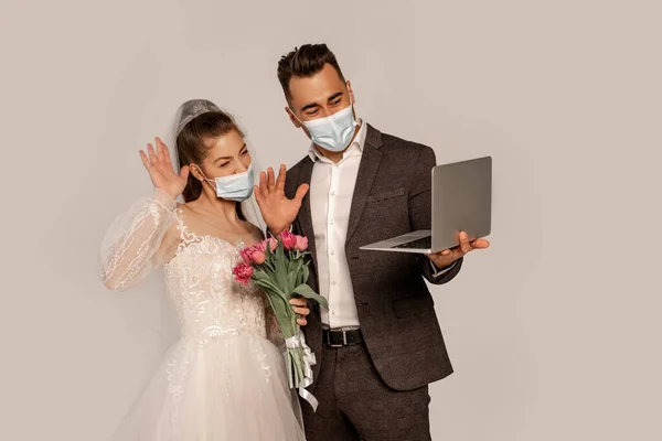 Bride and groom in medical masks waving hands during video call isolated on grey — Stock Photo