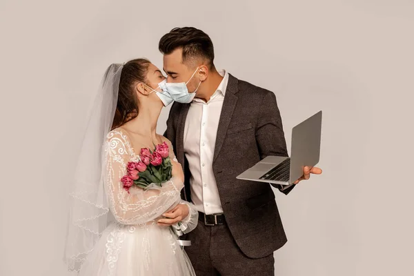 Мужчина в медицинской маске целует невесту, держа ноутбук изолированным на серой — стоковое фото
