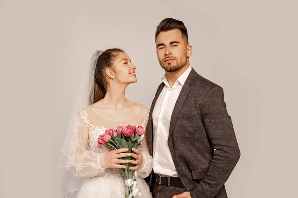 Happy bride looking at confident groom standing with hand in pocket isolated on grey — Fotografia de Stock