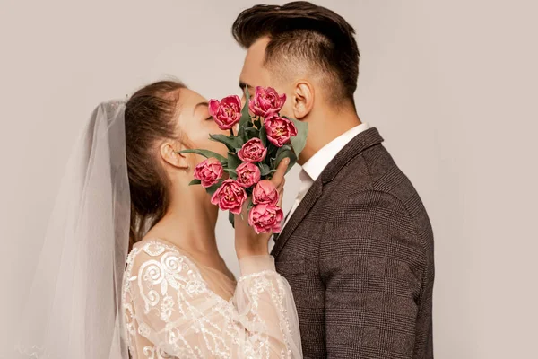 Молодые молодожены целуются, затемняя лица тюльпанами, изолированными на сером — стоковое фото