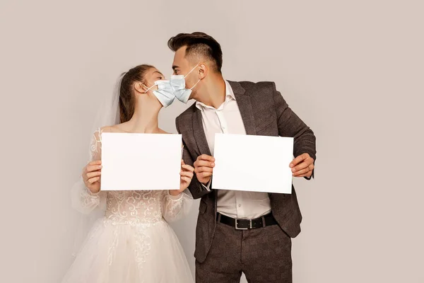 Newlyweds in safety masks holding blank cards while kissing isolated on grey — Stock Photo