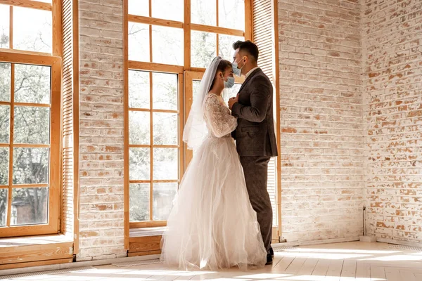 Full length view of young newlyweds in protective masks standing near large windows — Stock Photo