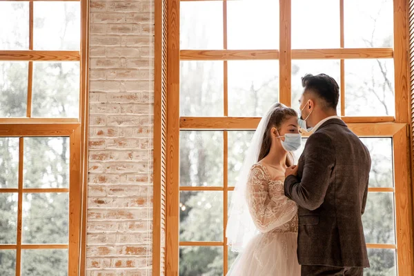 Newlyweds in protective masks near large windows at home — Stock Photo