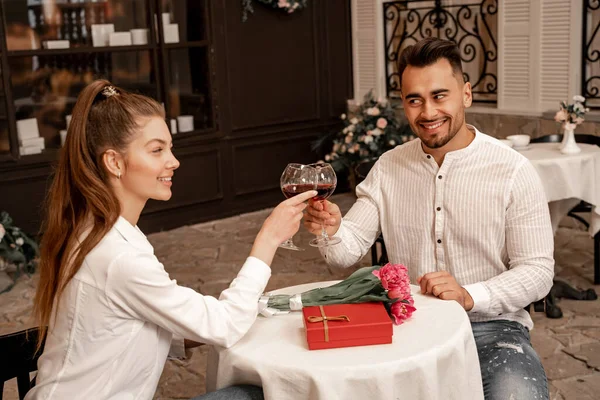 Happy couple clinking wine glasses near gift box and tulips in cafe — Photo de stock