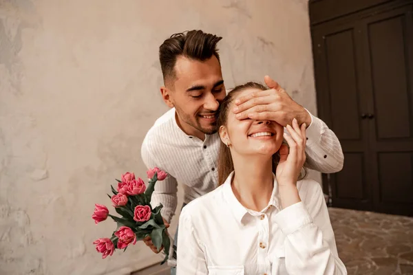 Улыбающийся мужчина, держащий букет тюльпанов, закрывая руками глаза счастливой подружки — стоковое фото