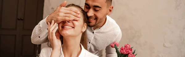 Glücklicher Mann, der die Augen einer fröhlichen Freundin mit der Hand bedeckt, während er Blumen hält, Banner — Stockfoto
