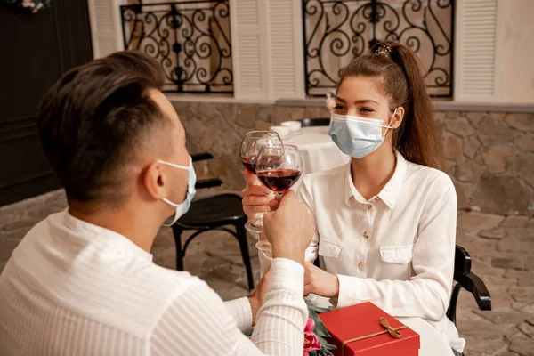 Young couple in medical masks clinking wine glasses during date in restaurant — Photo de stock