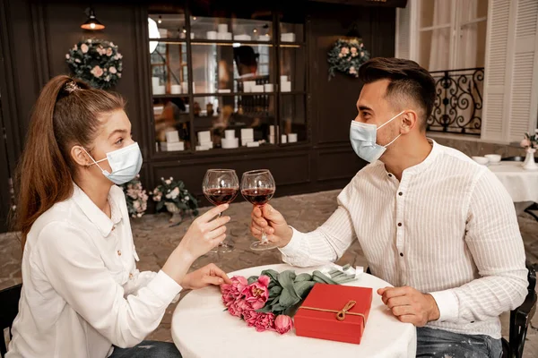 Man and woman in medical masks clinking wine glasses near gift box and tulips in restaurant — Fotografia de Stock