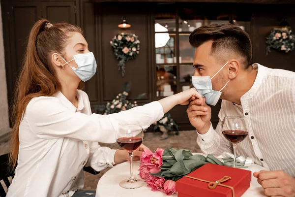 Young man in protective mask kissing hand of woman during date in cafe — Photo de stock